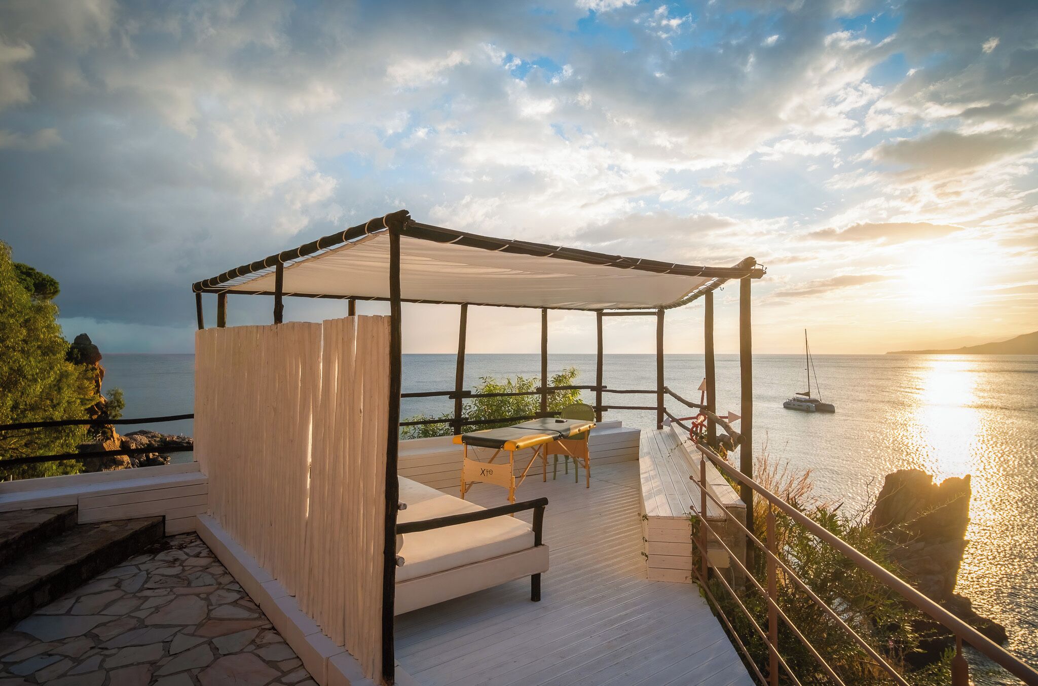 A covered outdoor seating area with a table and chairs, overlooking a body of water and a sunset.
