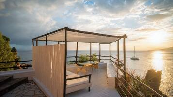 A covered outdoor seating area with a table and chairs, overlooking a body of water and a sunset.