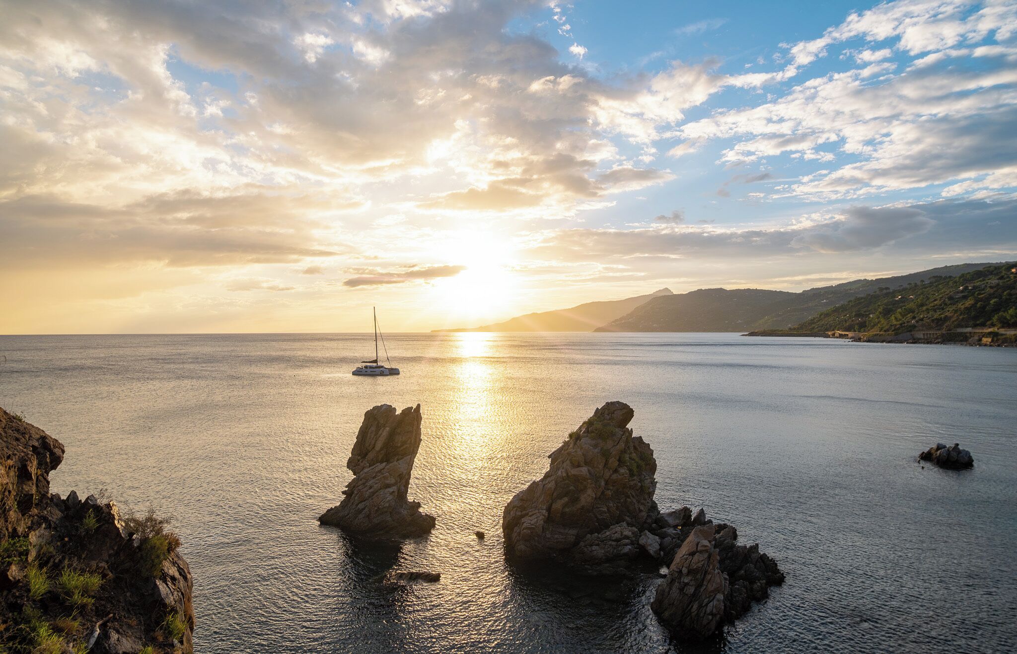 A coastal scene with a sailboat on the water, rocky formations, and a sunset.