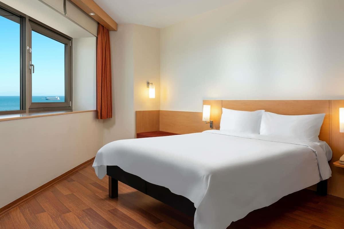 A hotel room with a large bed, wooden headboard, two bedside lamps, a window with curtains, and a view of the sea.