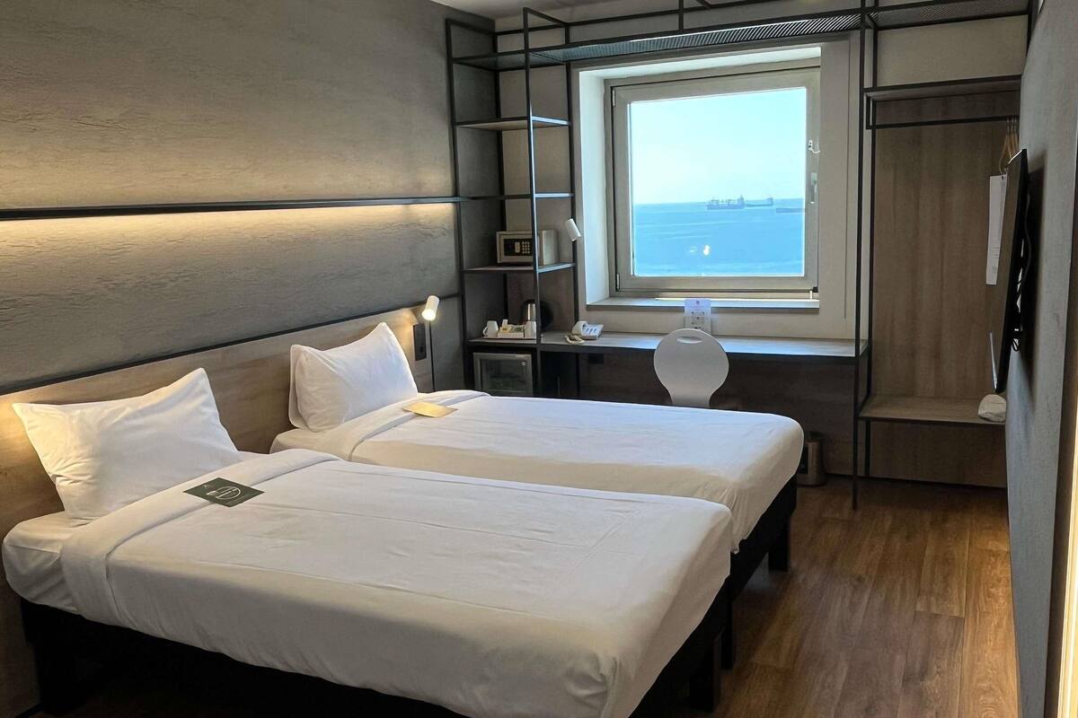 A hotel room with two beds, a desk, a chair, a small table, a window with a view of the sea, and a wardrobe.