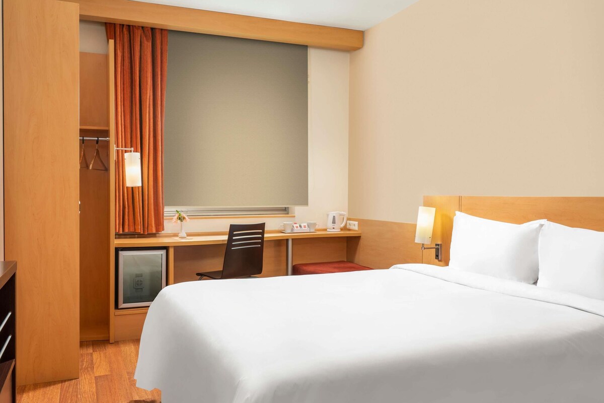 A hotel room with a bed, desk, chair, and a small kitchenette area.