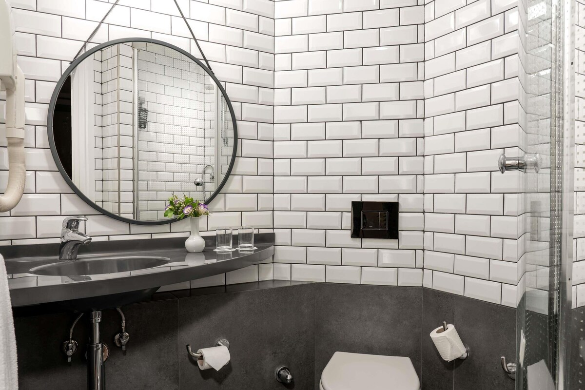 A modern bathroom with white subway tiles, a round mirror, a dark countertop, a toilet, and a shower area.
