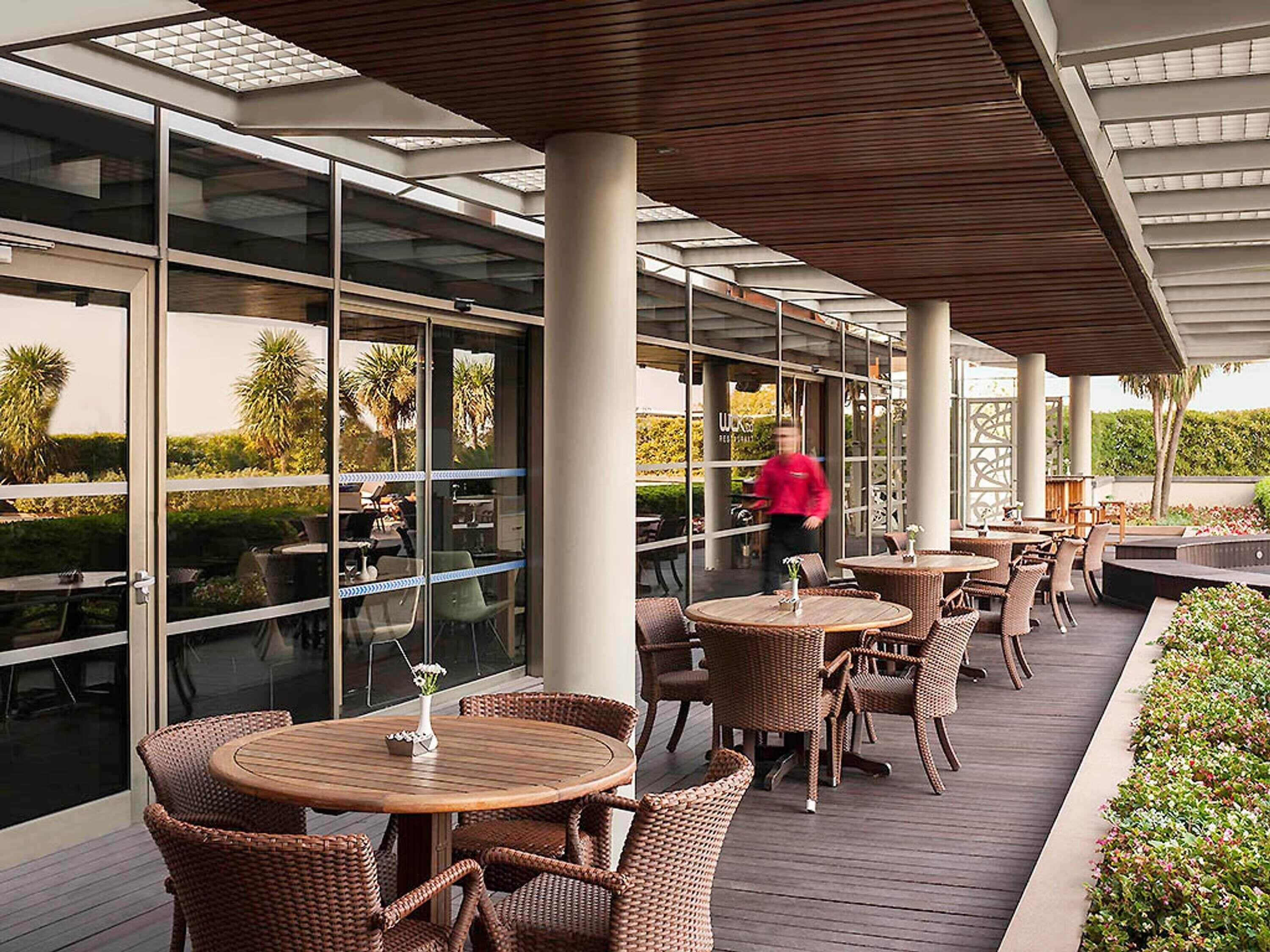 An outdoor patio area with round wooden tables and wicker chairs, large glass windows, and a view of palm trees.