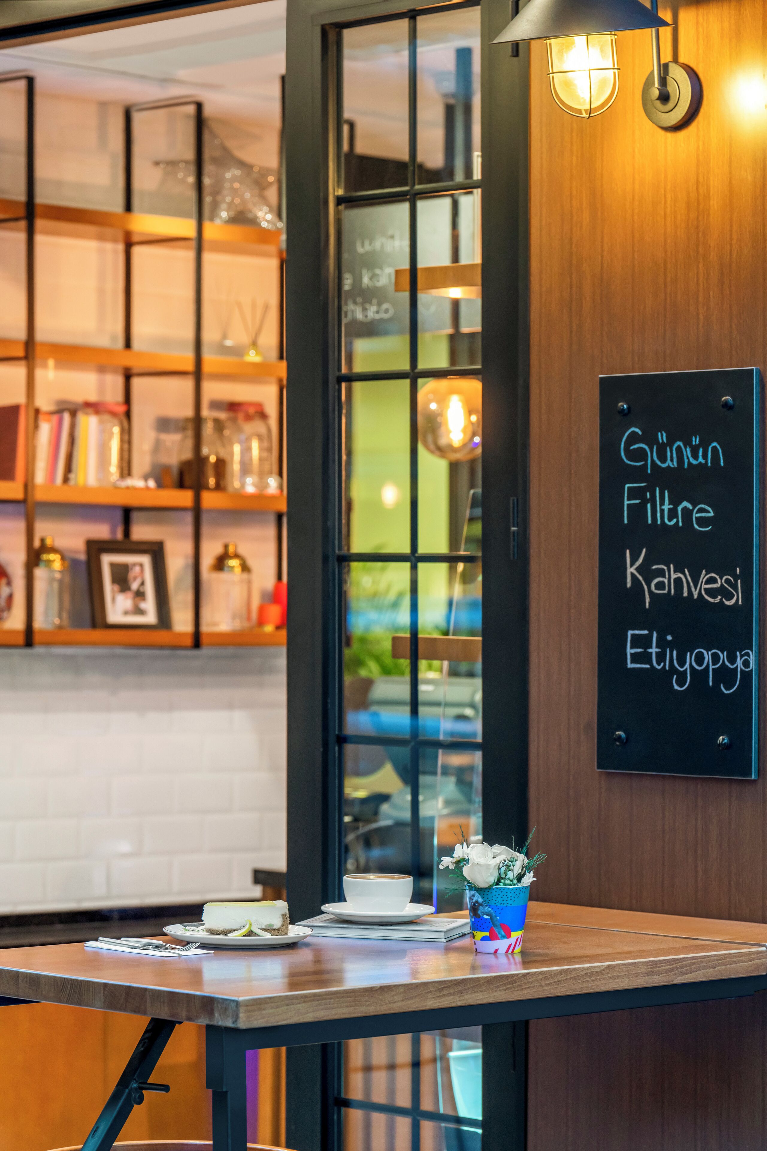 Een café-interieur met een houten tafel, een krijtbordmenu en planken met boeken en potjes.