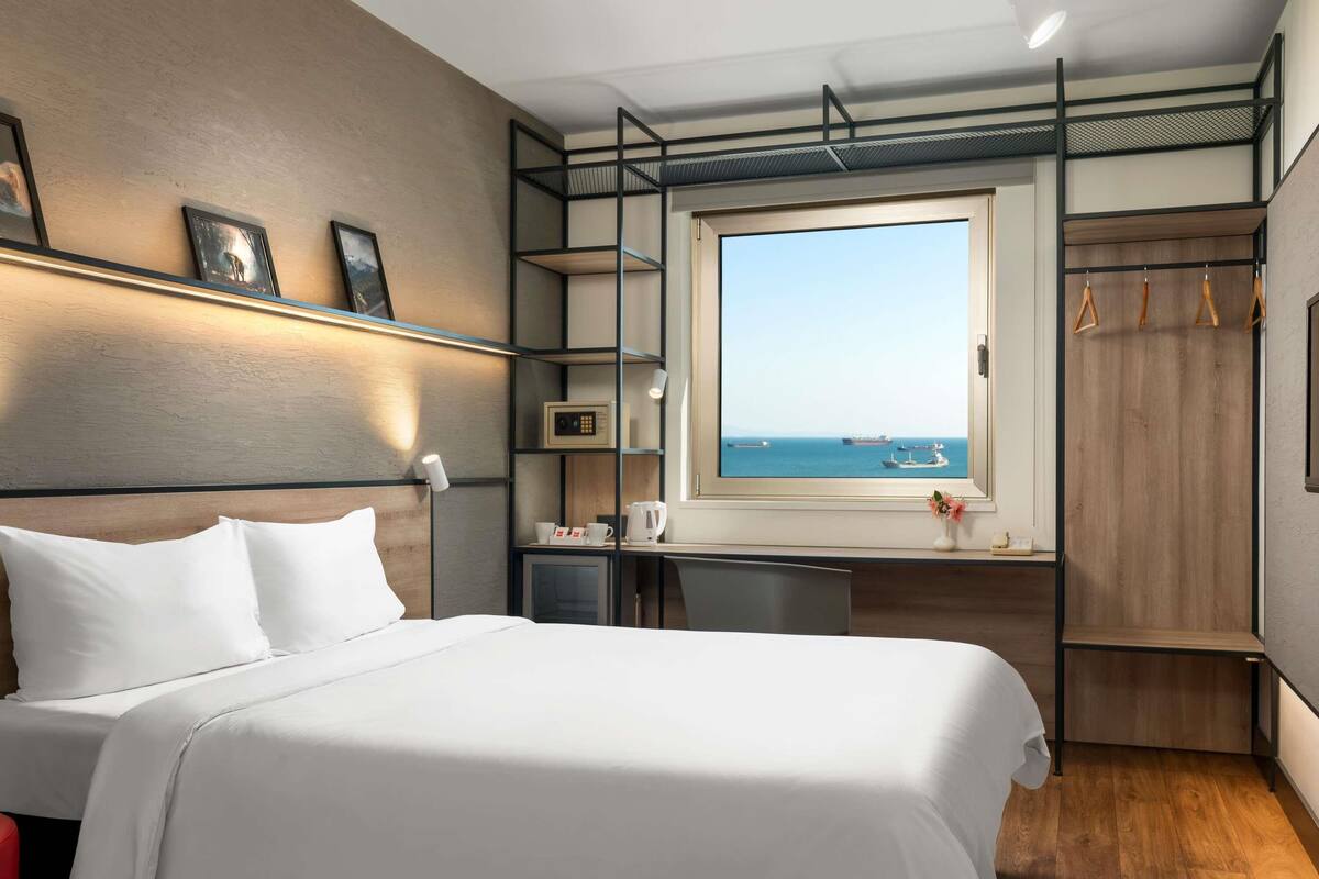A modern hotel room with a bed, a desk, a wardrobe, and a window with a view of the sea.