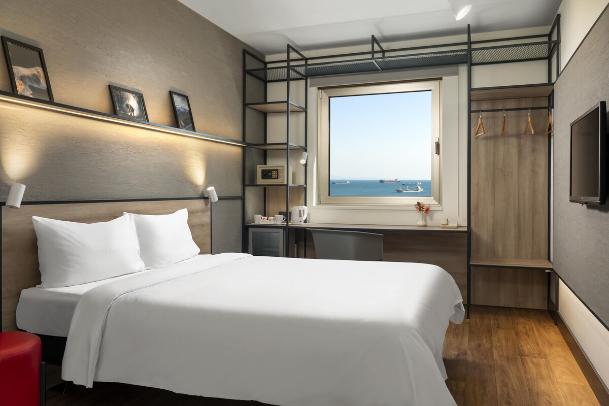 A modern hotel room with a large bed, a desk, a TV, and a window with a view of the sea.