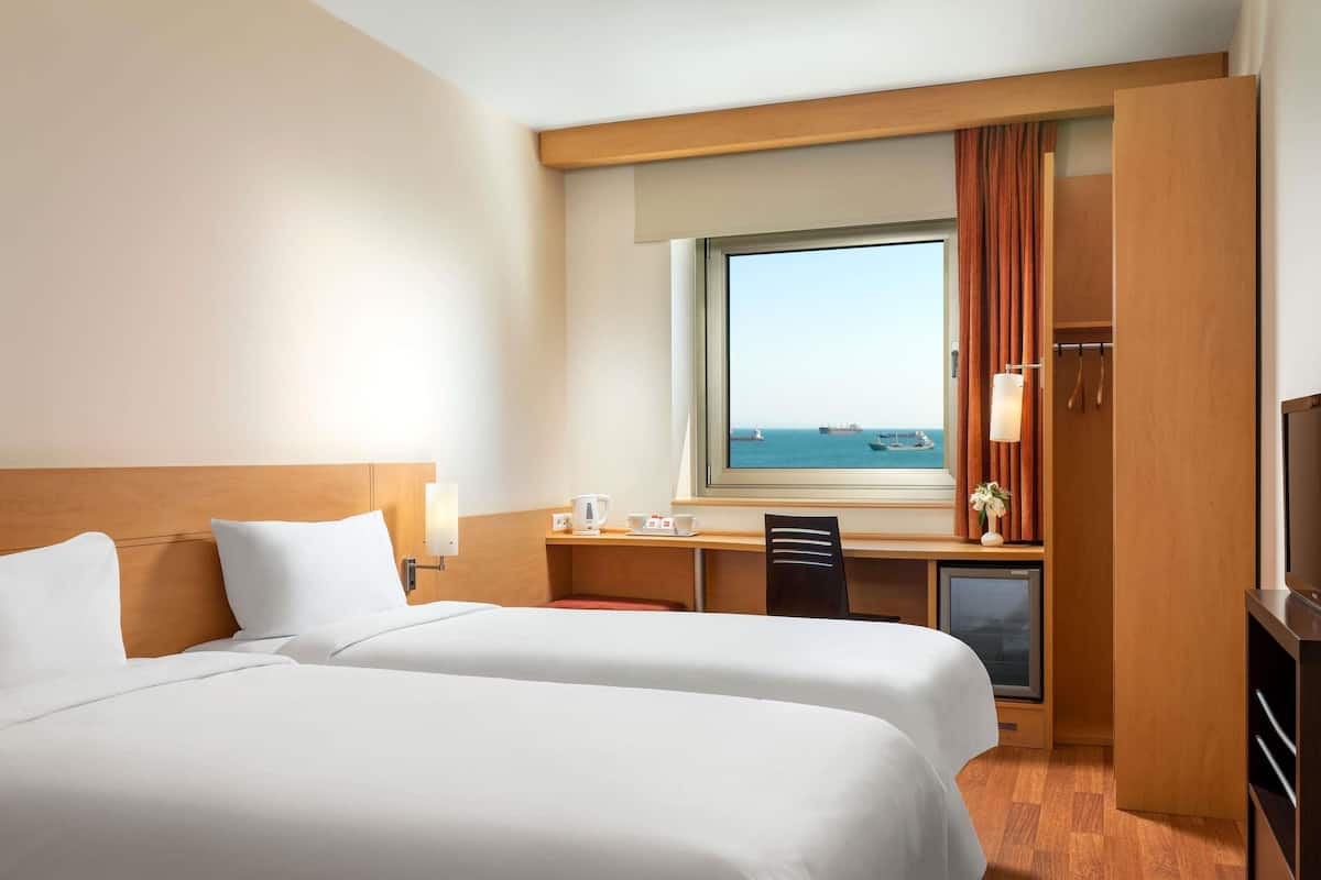 A hotel room with two beds, a desk with a computer, a TV, and a window with a view of the sea.