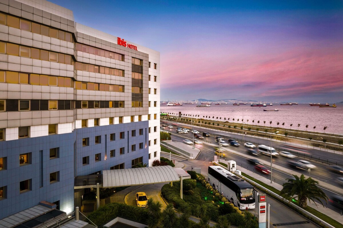 A modern hotel with a prominent "ibis hotel" sign, a waterfront view, and a busy road with vehicles.