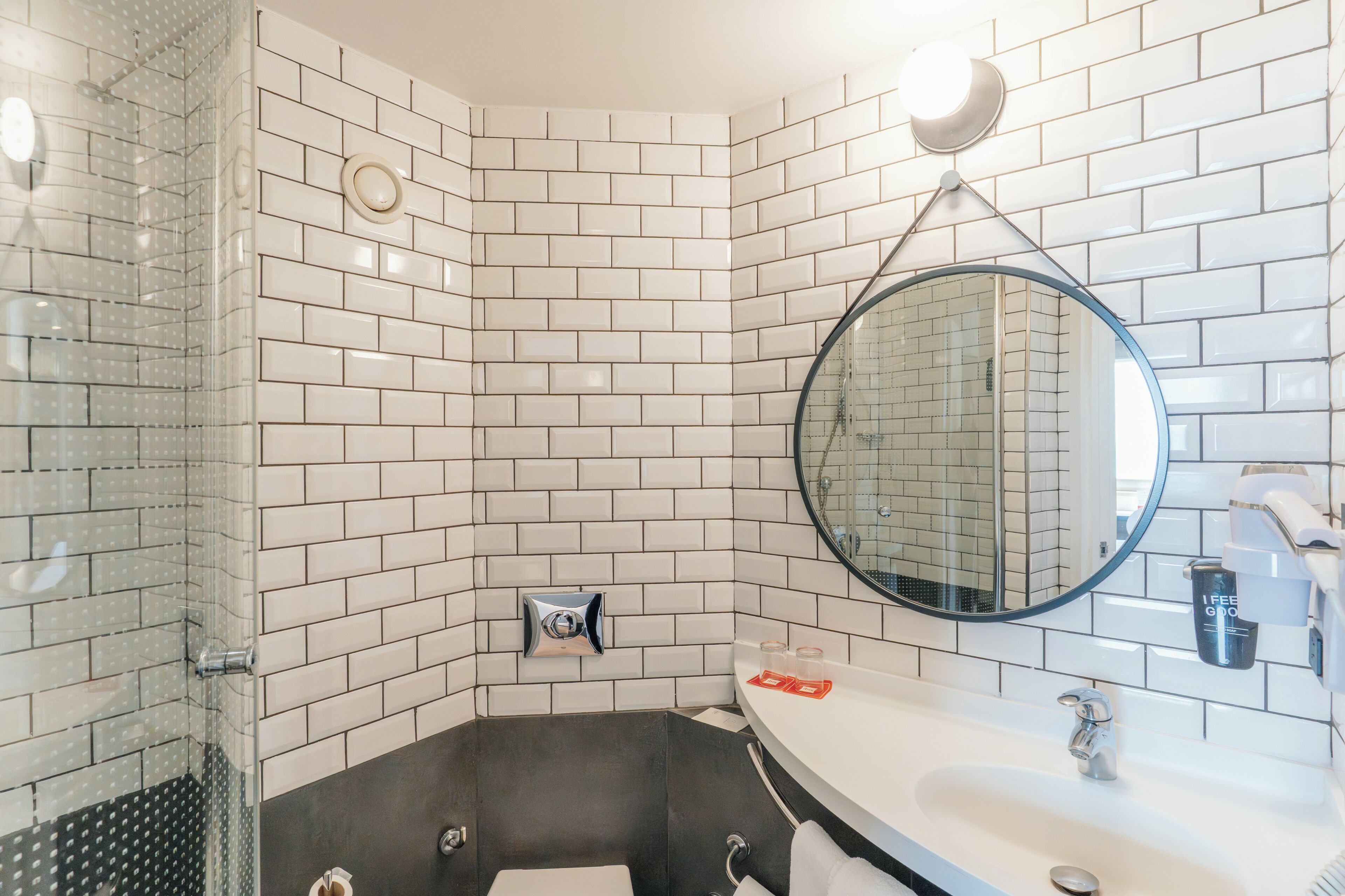 Ein modernes Badezimmer mit weißen, rechteckigen Fliesen, einer Duschkabine aus Glas, einem runden Spiegel, einer schwarzen Badewanne und einem weißen Waschbecken mit Wasserhahn.