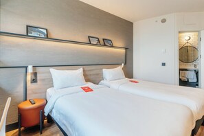 A hotel room with two beds, a small orange stool, a remote control, and a wall-mounted shelf with framed pictures. - Ibis Istanbul Zeytinburnu (Istanbul)