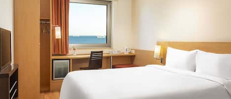 A hotel room with a bed, desk, chair, and a view of the sea through a window.