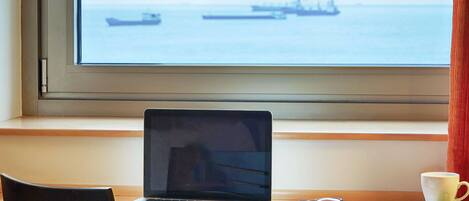 A laptop on a wooden desk by a window with a view of the sea and ships.