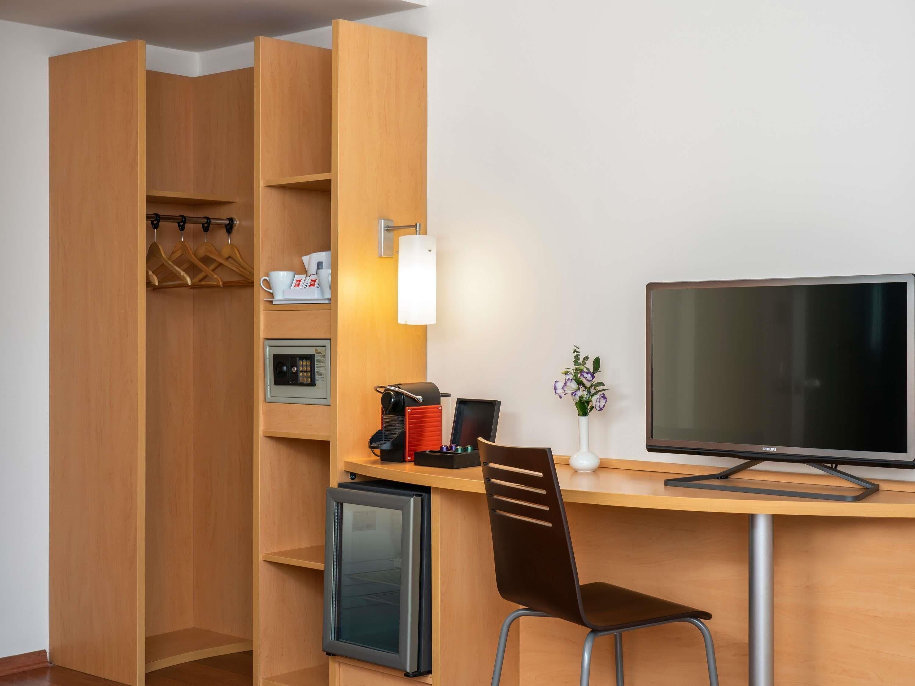 A compact hotel room with a wooden desk, a television, a coffee machine, a mini-fridge, and a seating area.