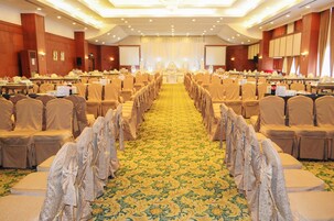 Un salón de banquetes con filas de mesas y sillas, piso alfombrado y un escenario al frente.