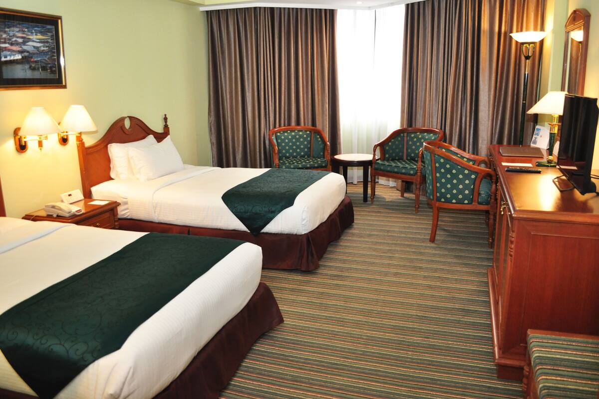 A hotel room with two beds, a desk, two chairs, a lamp, and a television.