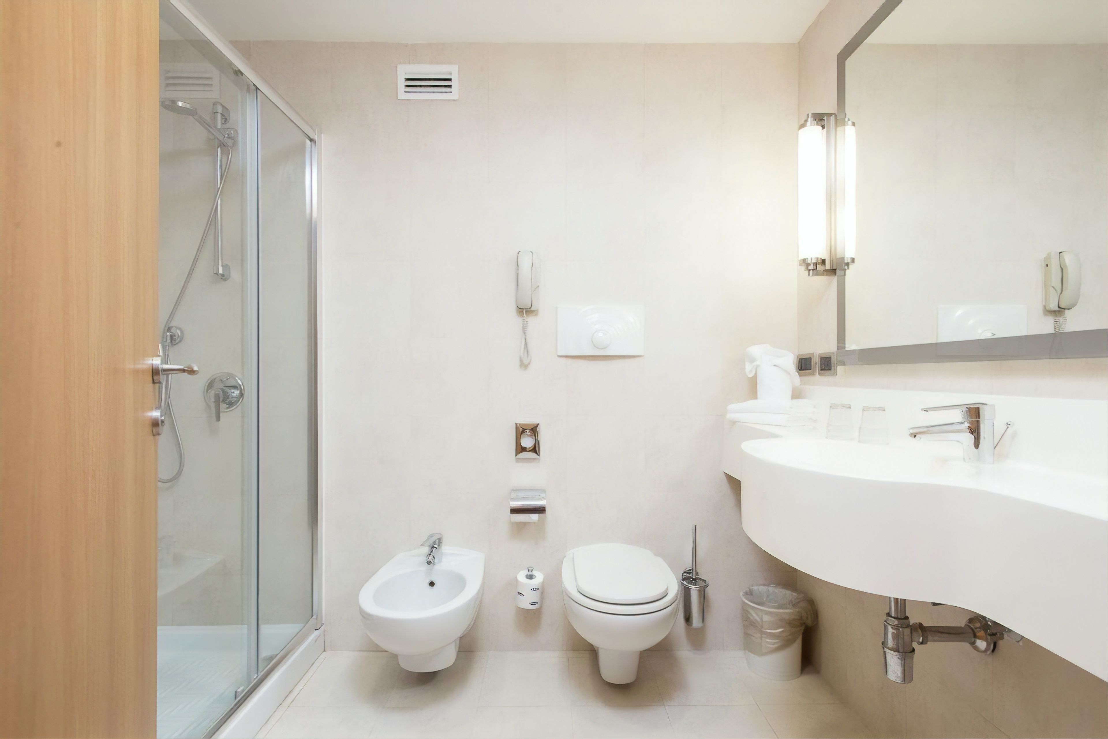 A modern bathroom with a glass shower enclosure, a bidet, a toilet, a sink with a mirror, and a trash bin.