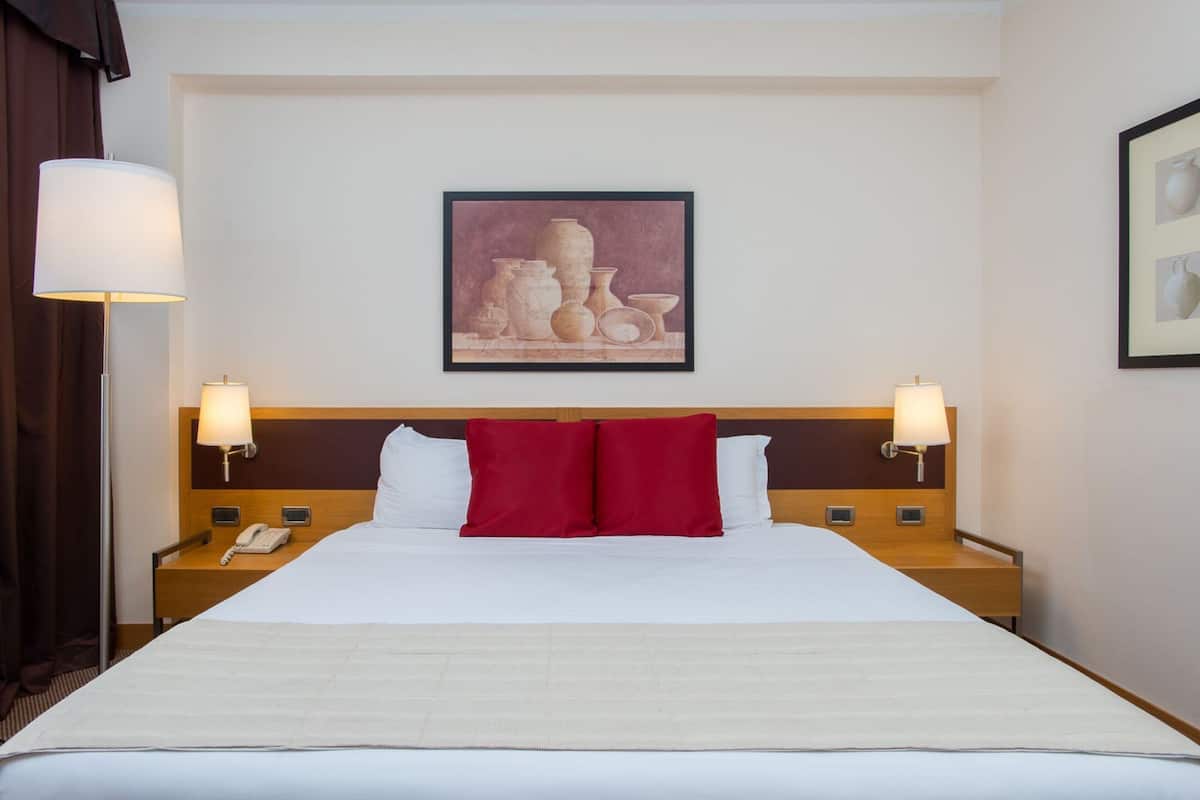 Habitación de hotel con una cama grande, dos mesitas de noche, una lámpara y una obra de arte enmarcada en la pared.