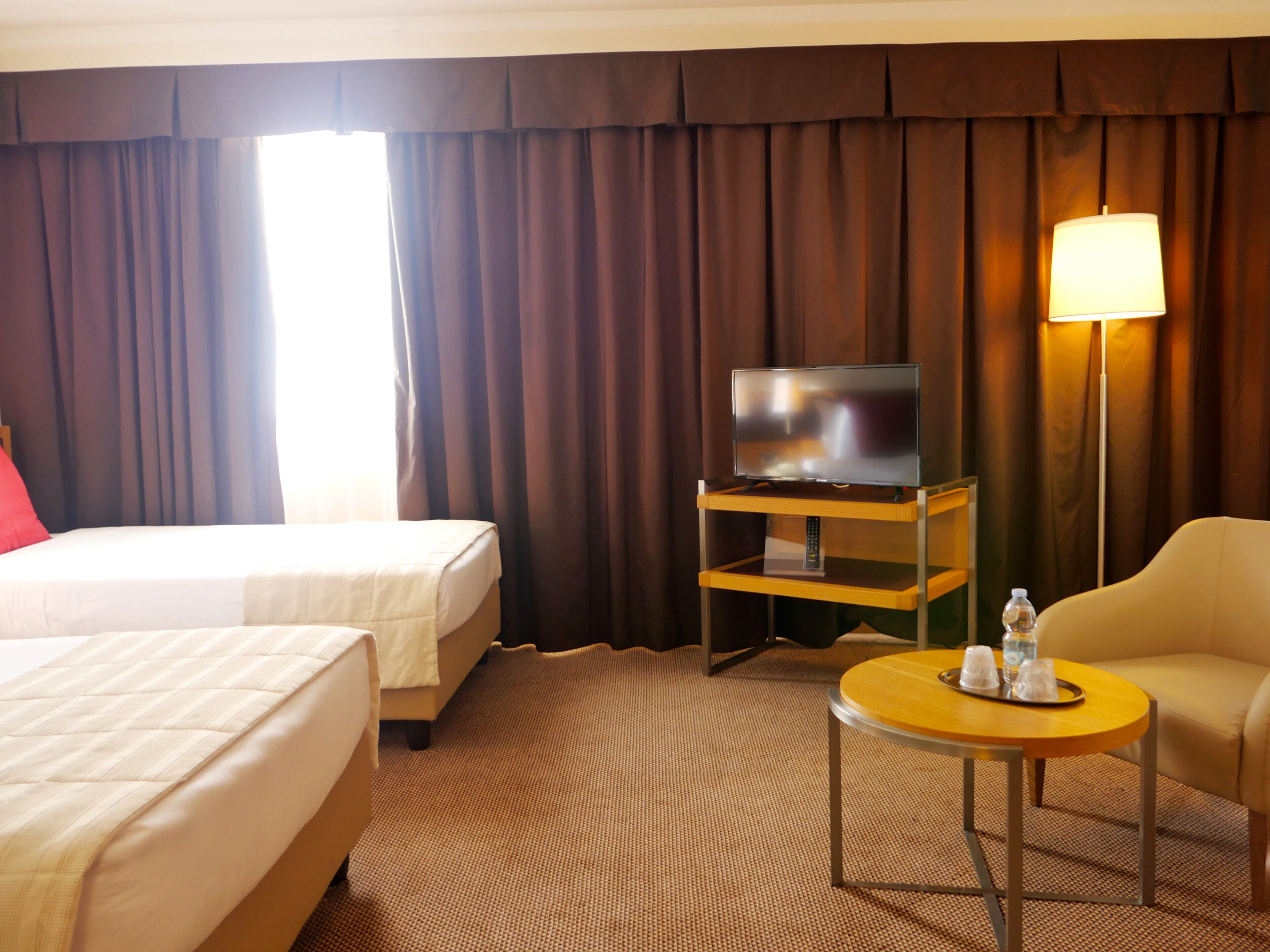 A hotel room with a bed, a TV on a stand, a lamp, a small table, and a chair.