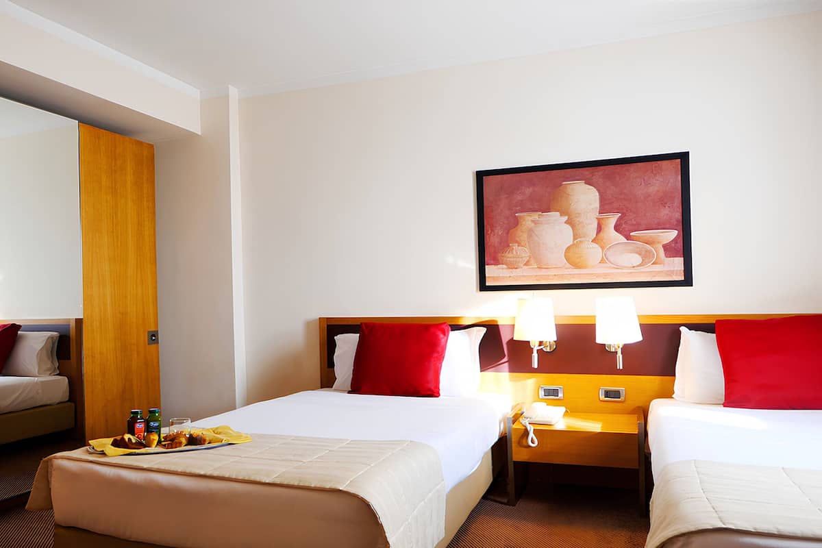 Una habitación de hotel con dos camas, una puerta de madera, un cuadro de cerámica y una mesita de noche con una lámpara.