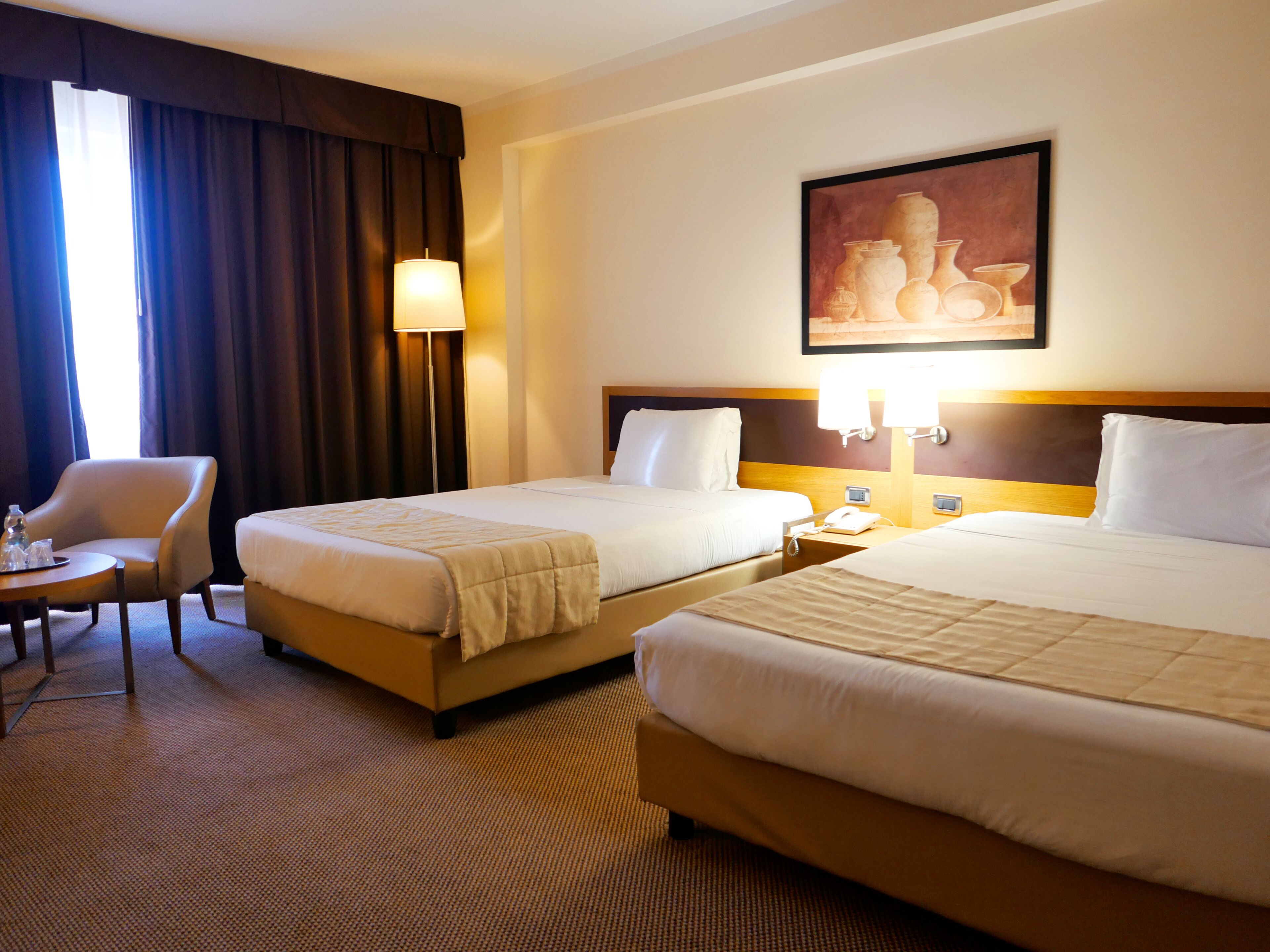 A hotel room with two beds, a chair, a lamp, and a painting on the wall.