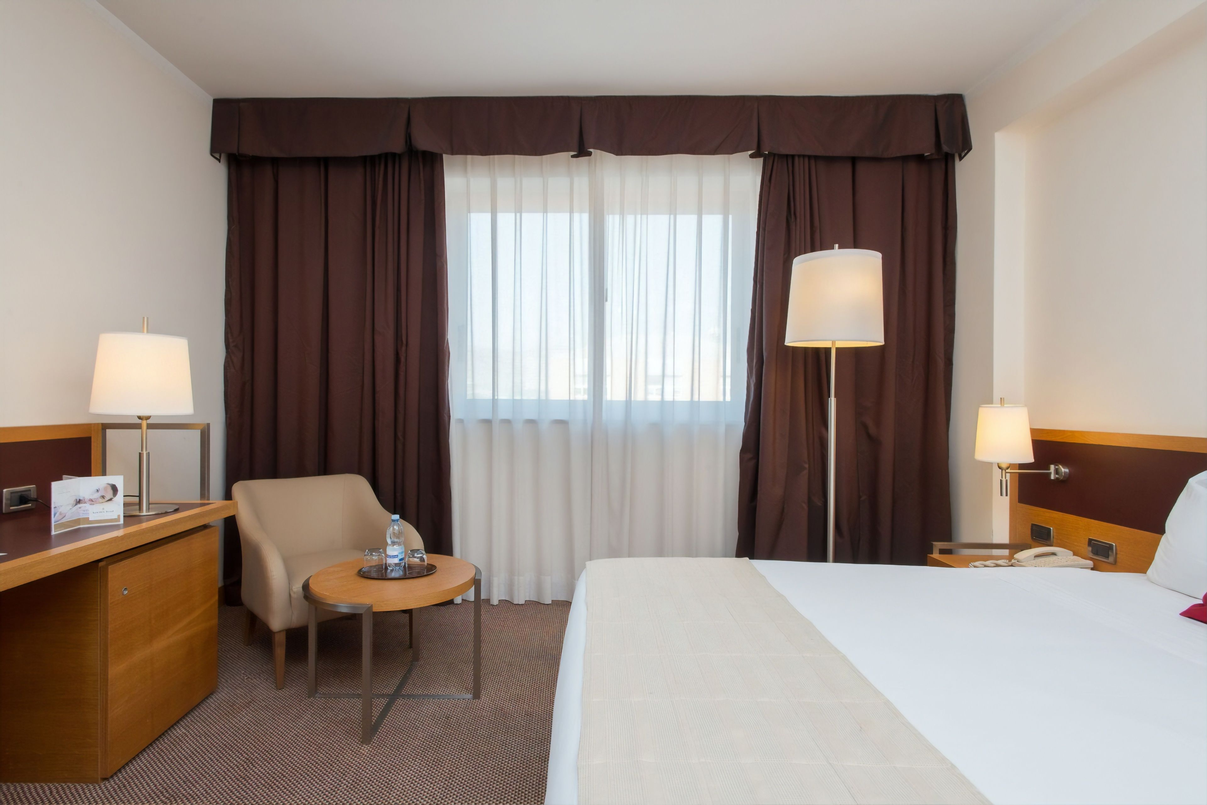 Una habitación de hotel con cama, escritorio, silla y dos lámparas.