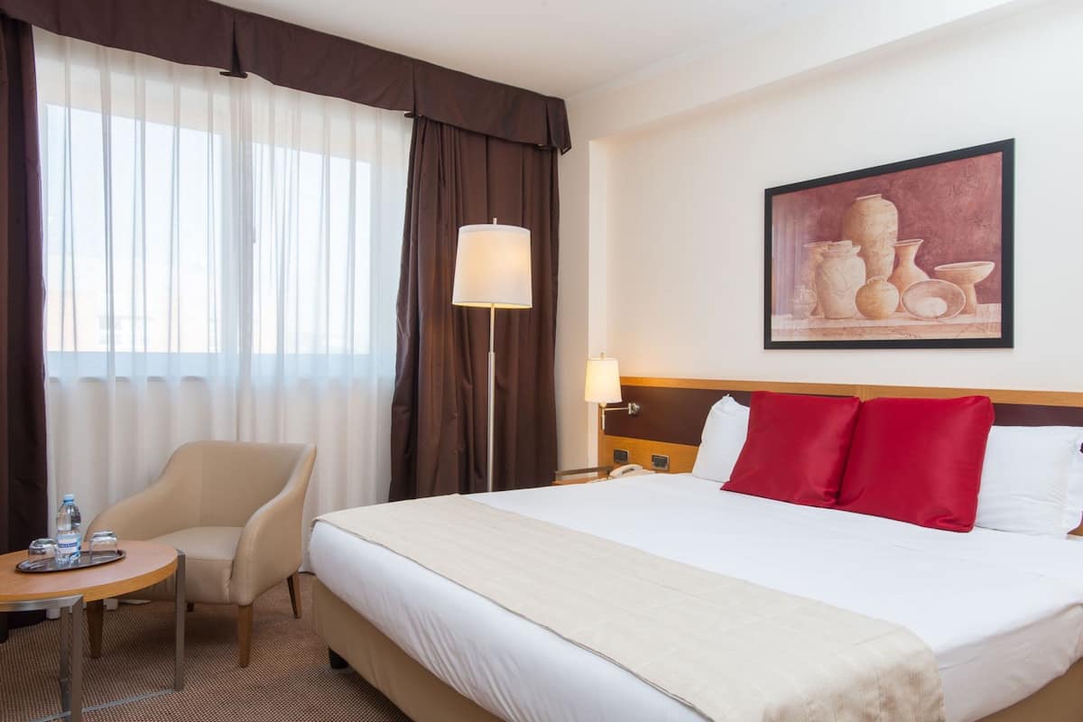 Habitación de hotel con cama, una silla, una mesa con bandeja, una lámpara y un cuadro en la pared.