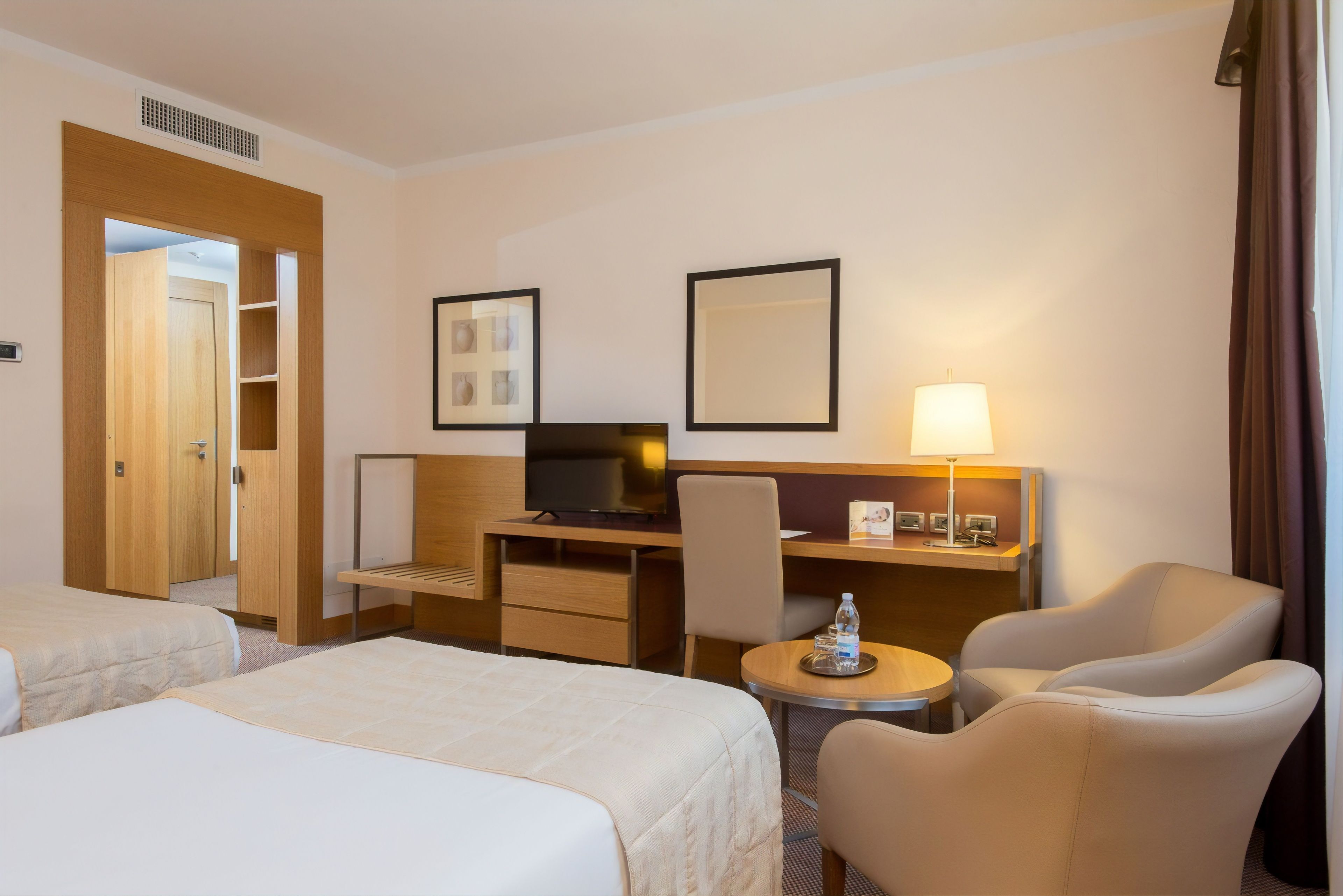 A hotel room with two beds, a desk with a chair, a television, a lamp, and a small table with a bottle.