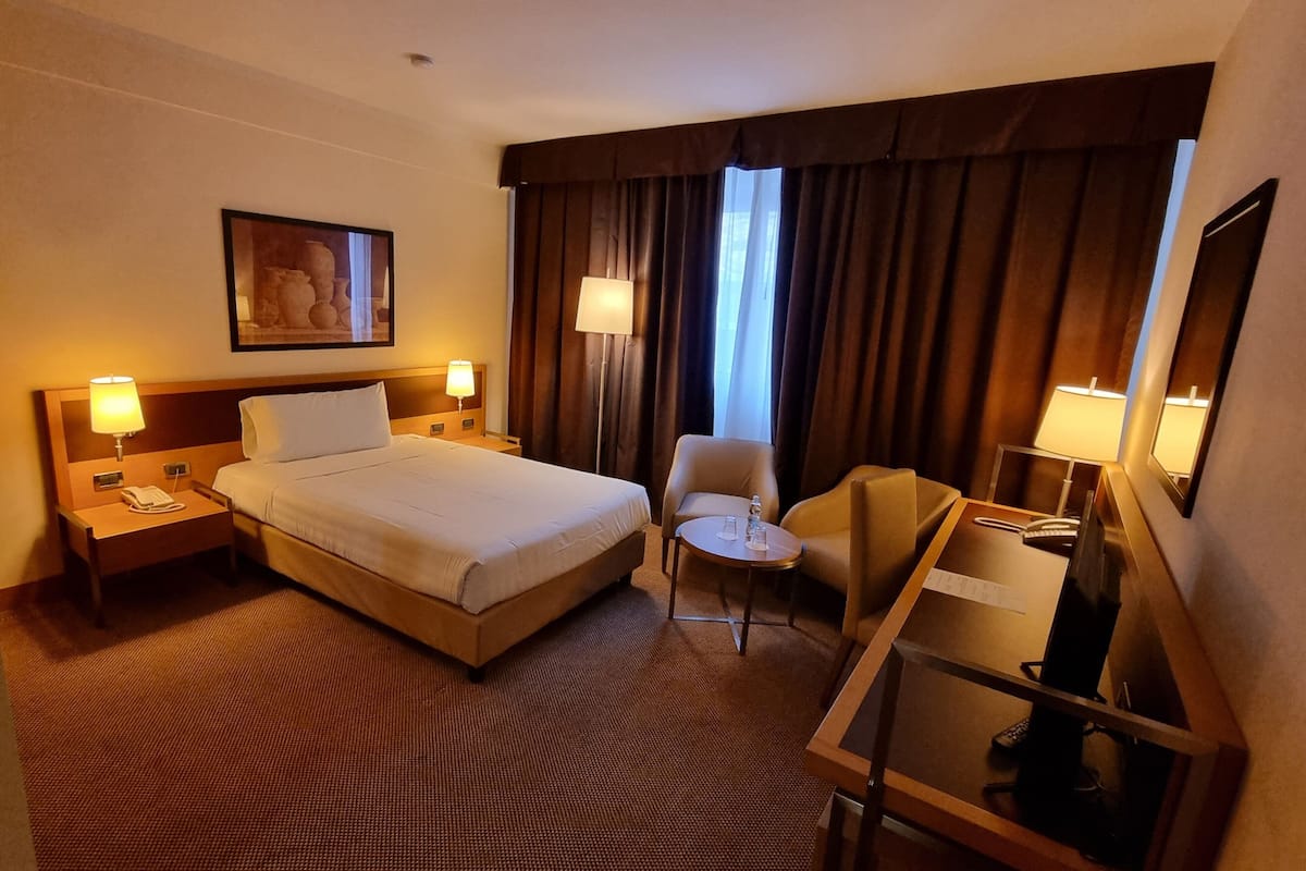 Una habitación de hotel con una cama grande, mesitas de noche con lámparas, una pequeña mesa redonda con una silla y un escritorio con ordenador y teléfono.