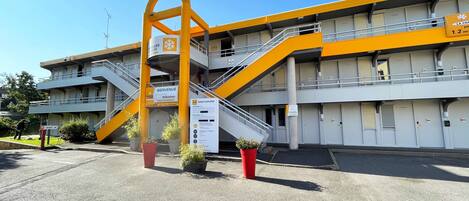 A multi-story hotel with a yellow and grey color scheme, featuring a central staircase, multiple balconies, and a welcoming entrance sign.