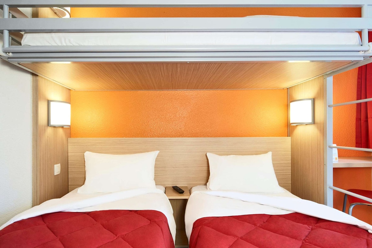 Habitación con literas, dos camas, cada una con colcha acolchada roja y almohadas blancas. Hay dos luces empotradas en la pared a cada lado de las camas, y un cabecero de madera.