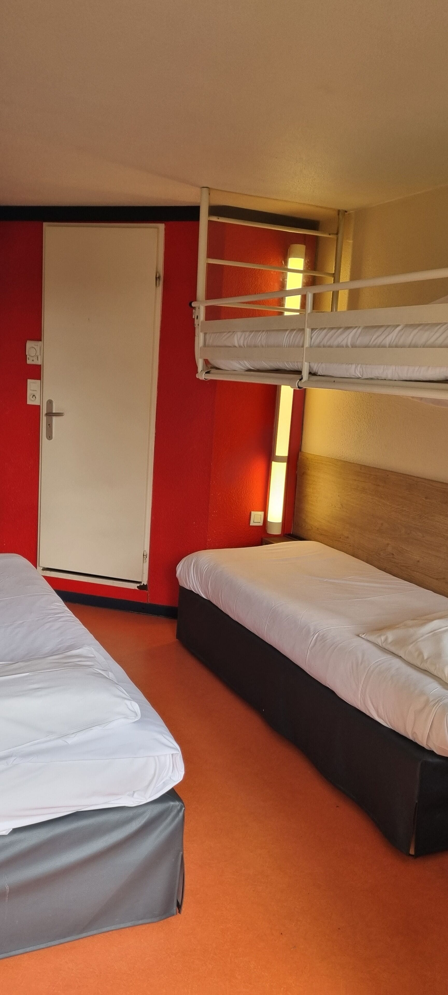 Una habitación con dos camas, una de ellas litera, y una puerta con interruptor de luz.