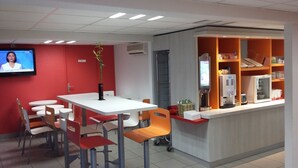 A modern cafe with a white table and orange chairs, a TV on the wall, and a vending machine. - Premiere Classe Rouen Sud - Oissel (Saint-Etienne-du-Rouvray)
