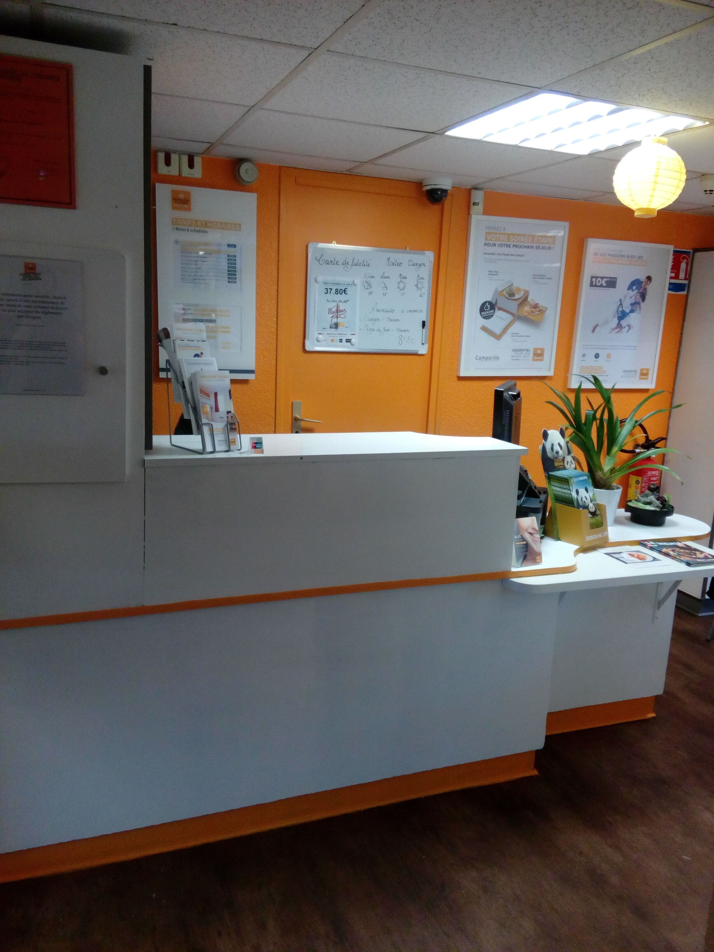 A reception desk with a white counter and orange accents, surrounded by informational posters and a security camera.