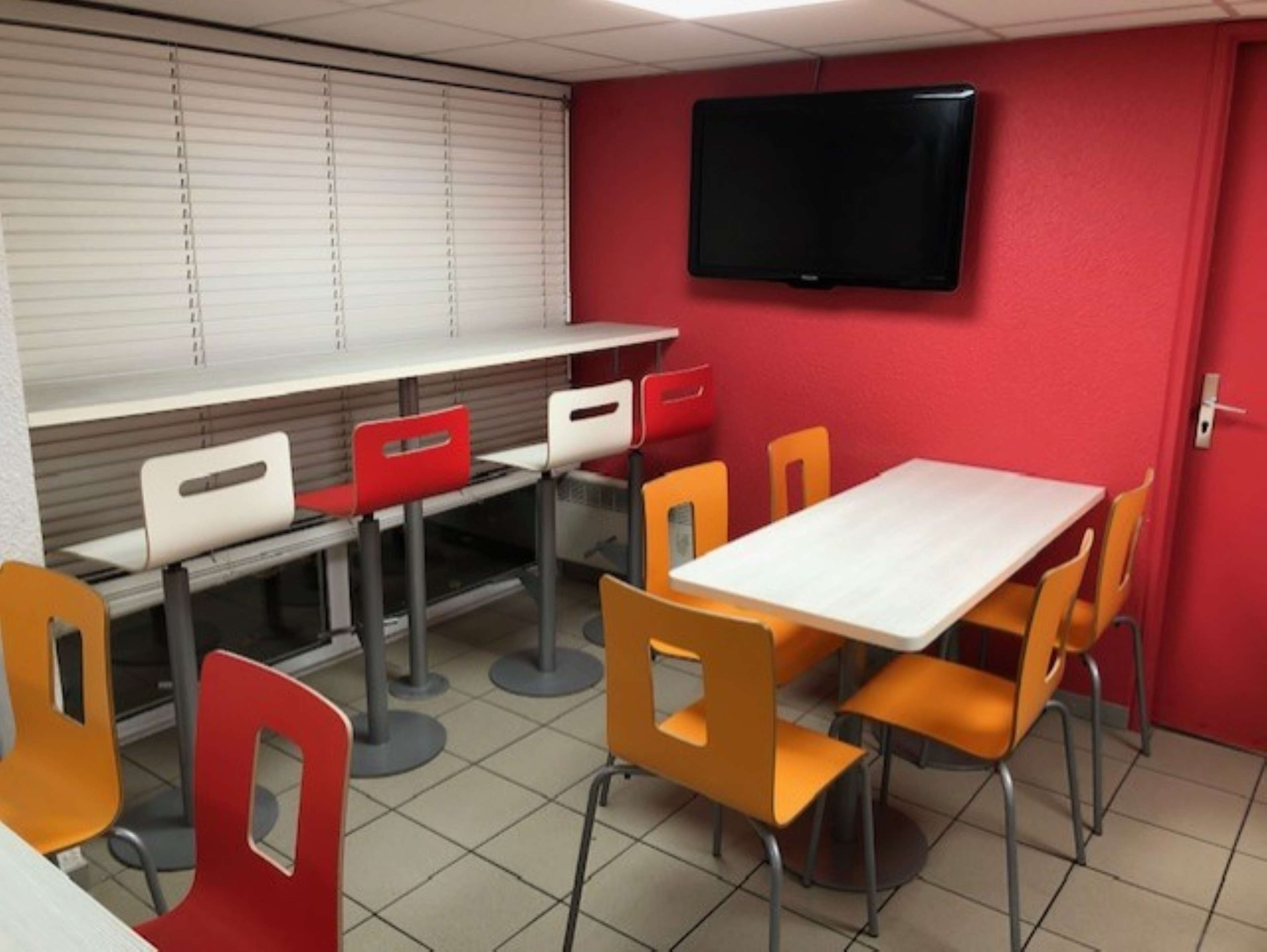 Un área de comedor con una mesa blanca y sillas naranjas, una pared roja y un televisor montado.