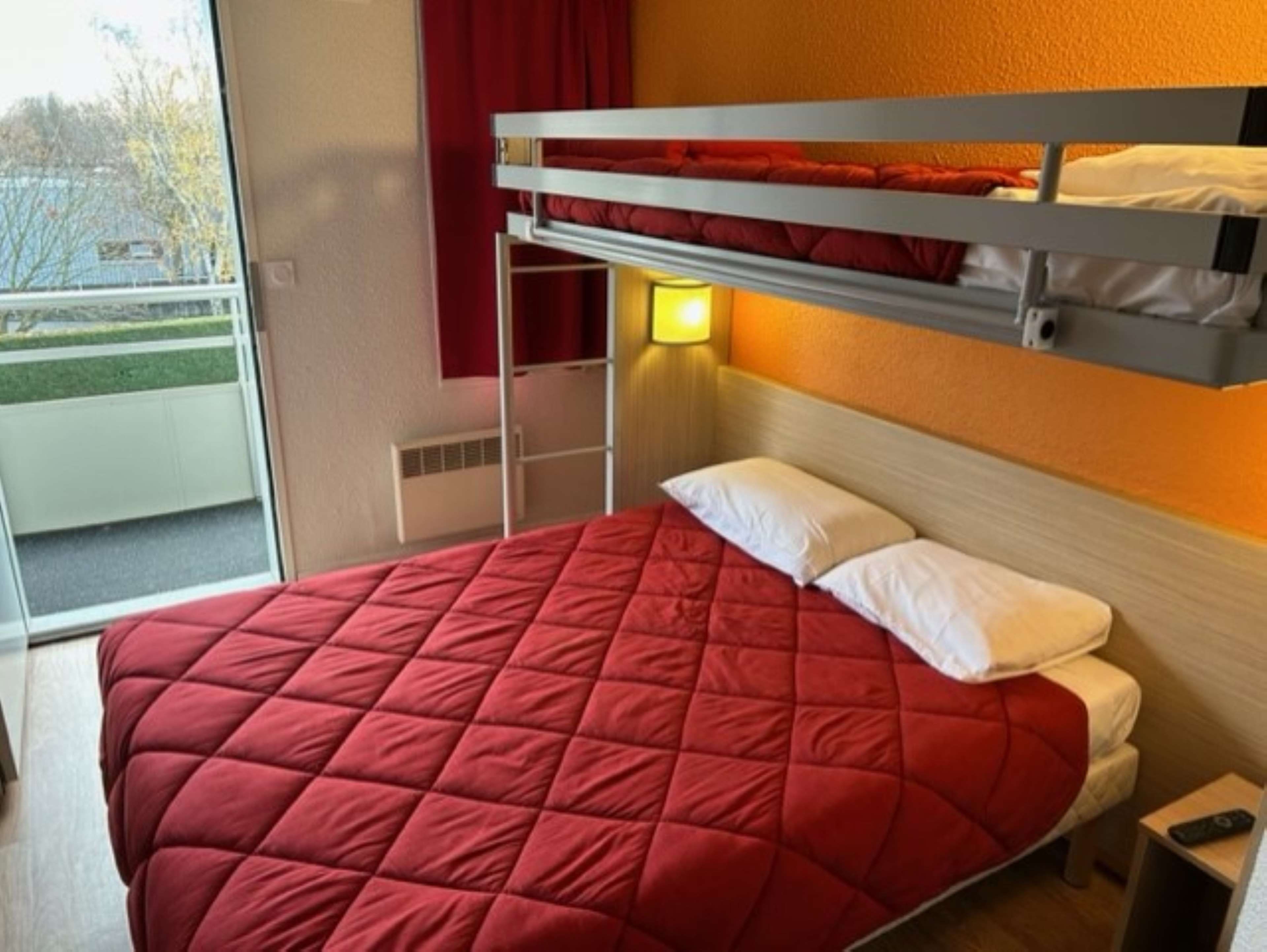 A bunk bed with a red quilted bedspread and white pillows, a bedside lamp, and a small bedside table with a remote control.