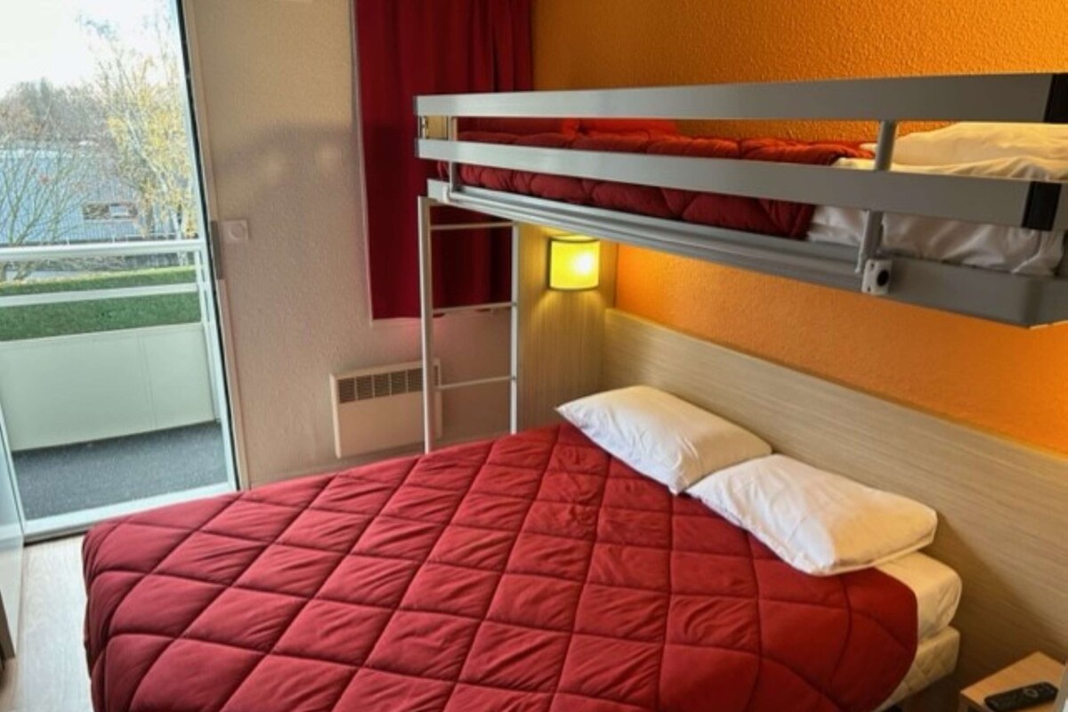 Ein Etagenbett mit roter gesteppter Bettdecke und weißen Kissen, eine Nachttischlampe und ein kleiner Tisch mit einer Fernbedienung.