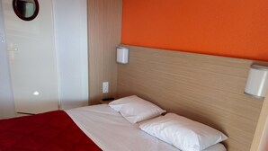 Una cama bien hecha con sábanas blancas y una manta roja, cabecera de madera y una mesita de noche con un control remoto.