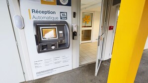 Un mostrador de recepción automatizado con pantalla digital, un ventanal pequeño y un cartel que indica los servicios del hotel.