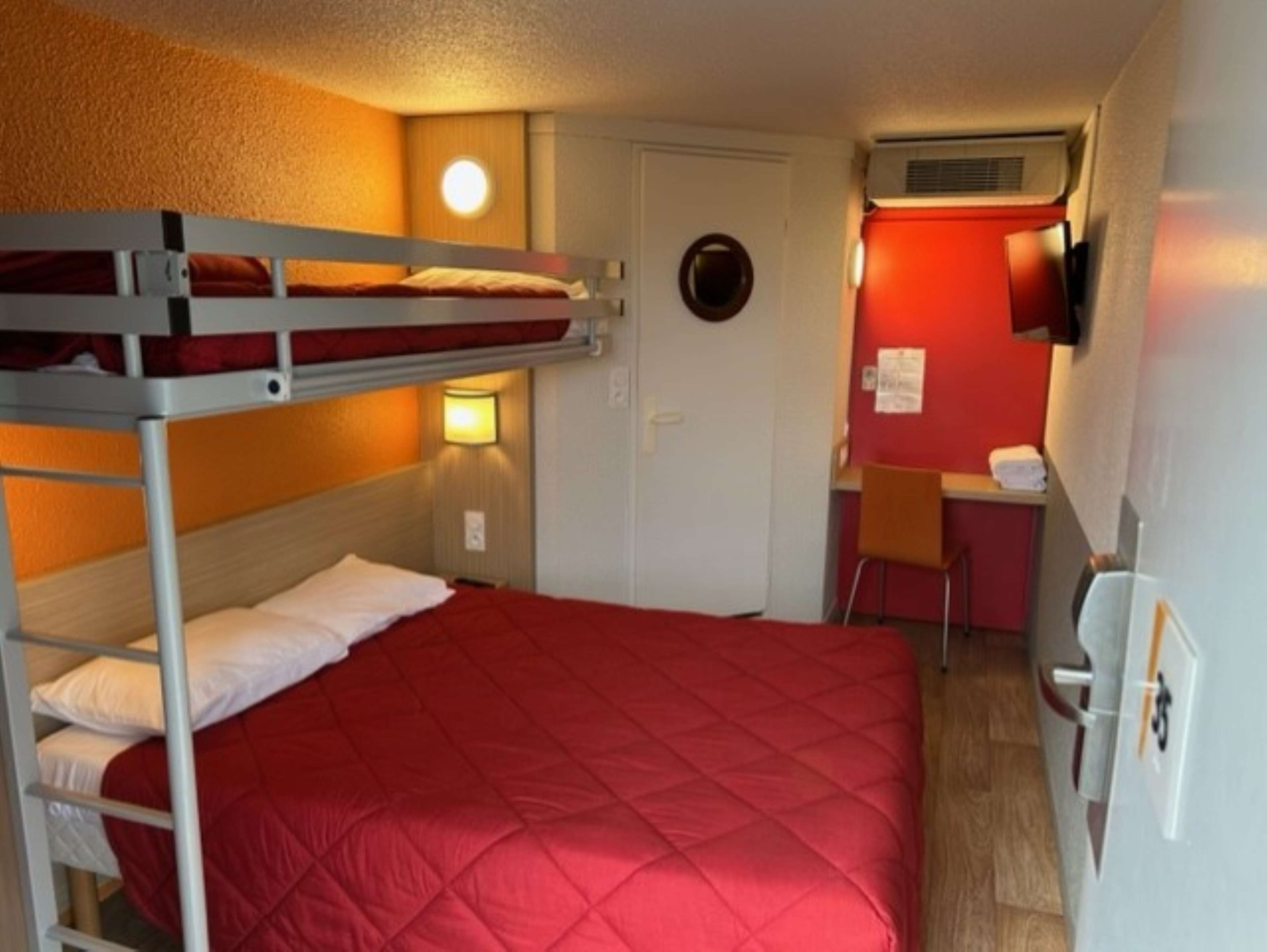 Habitación con literas, una colcha de patchwork roja, un escritorio con silla y un televisor montado en la pared.