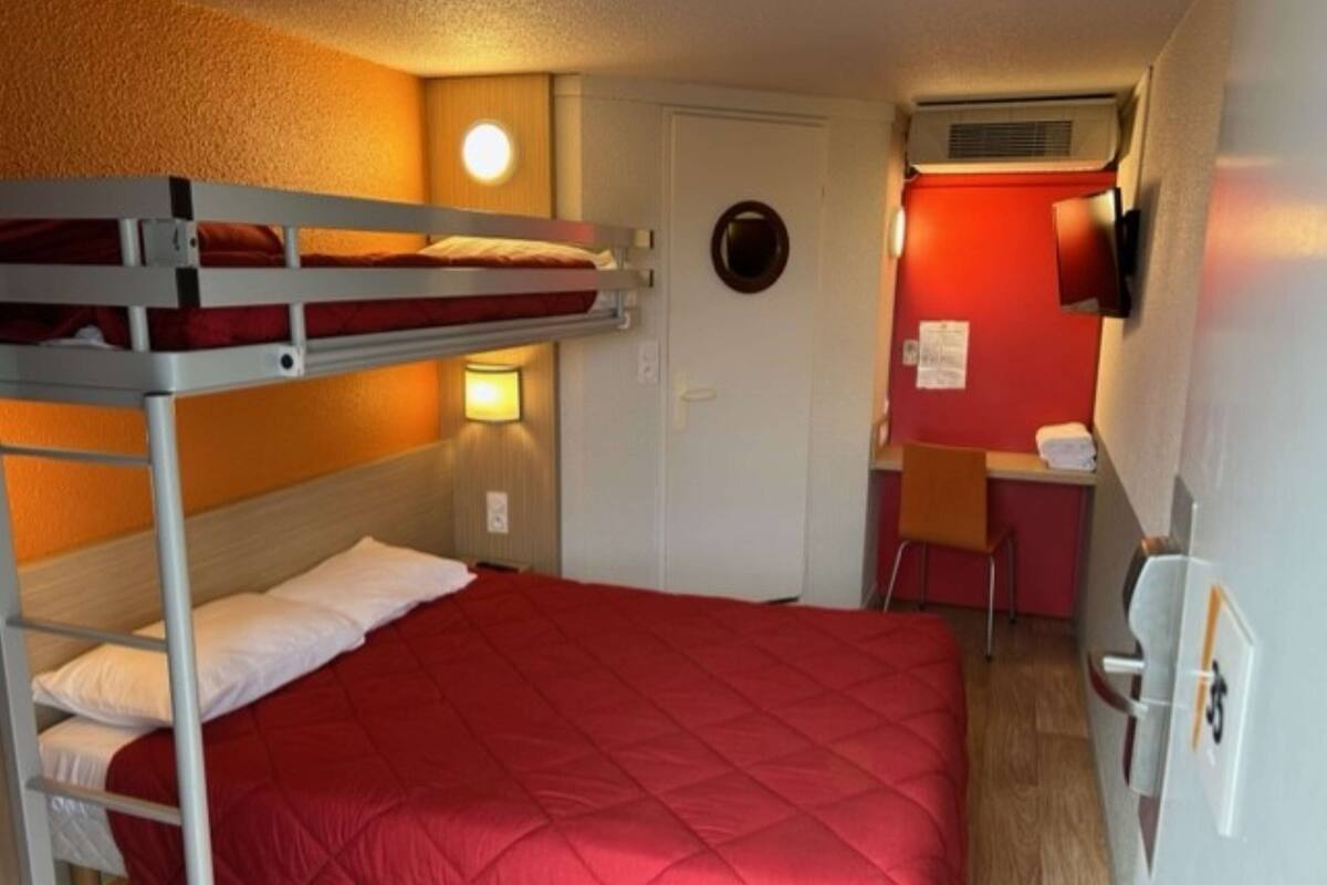A bunk bed room with a red quilted bedspread, a desk with a chair, and a wall-mounted TV.