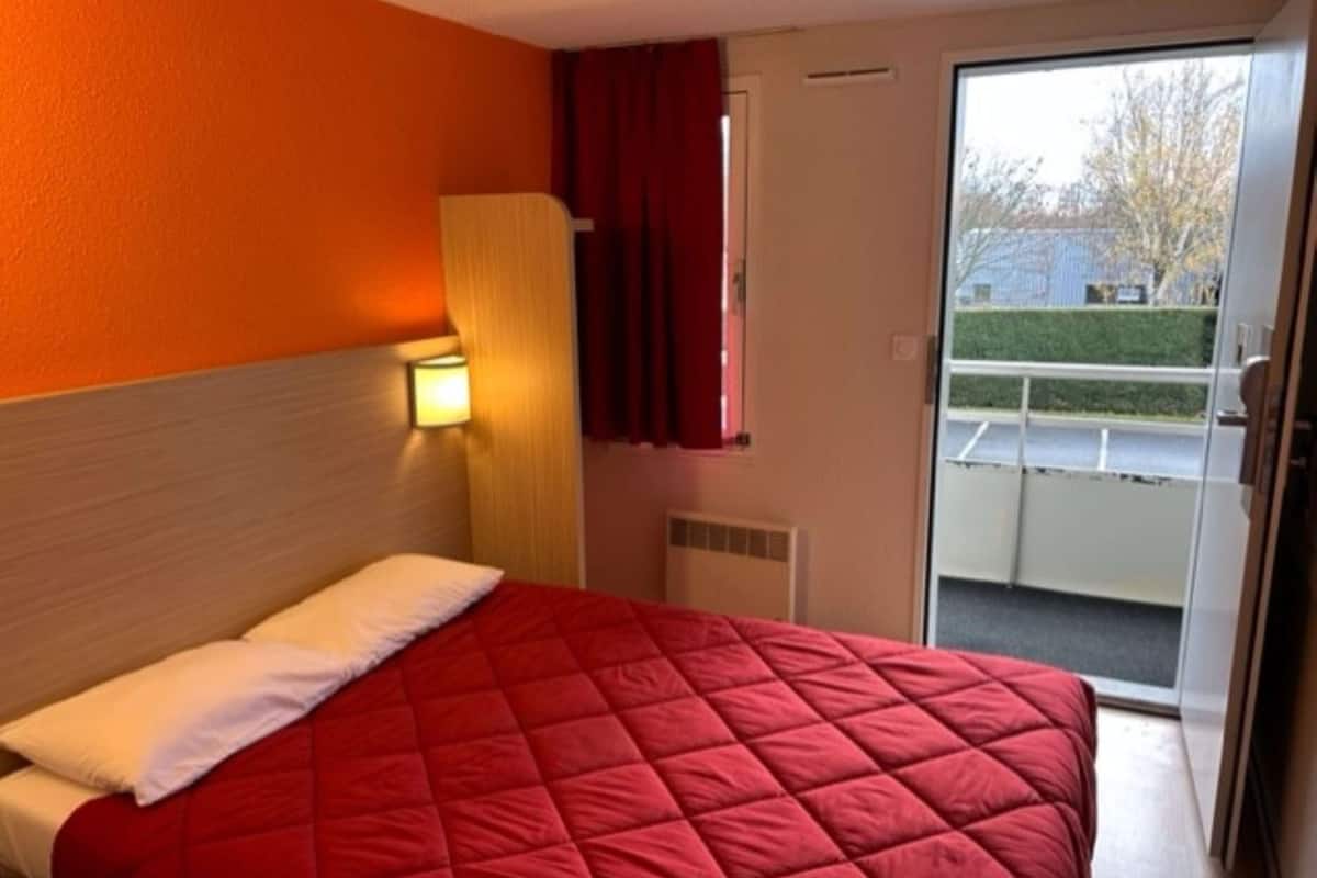 A hotel room with a bed, a nightstand, a window with curtains, and a view of the outside.
