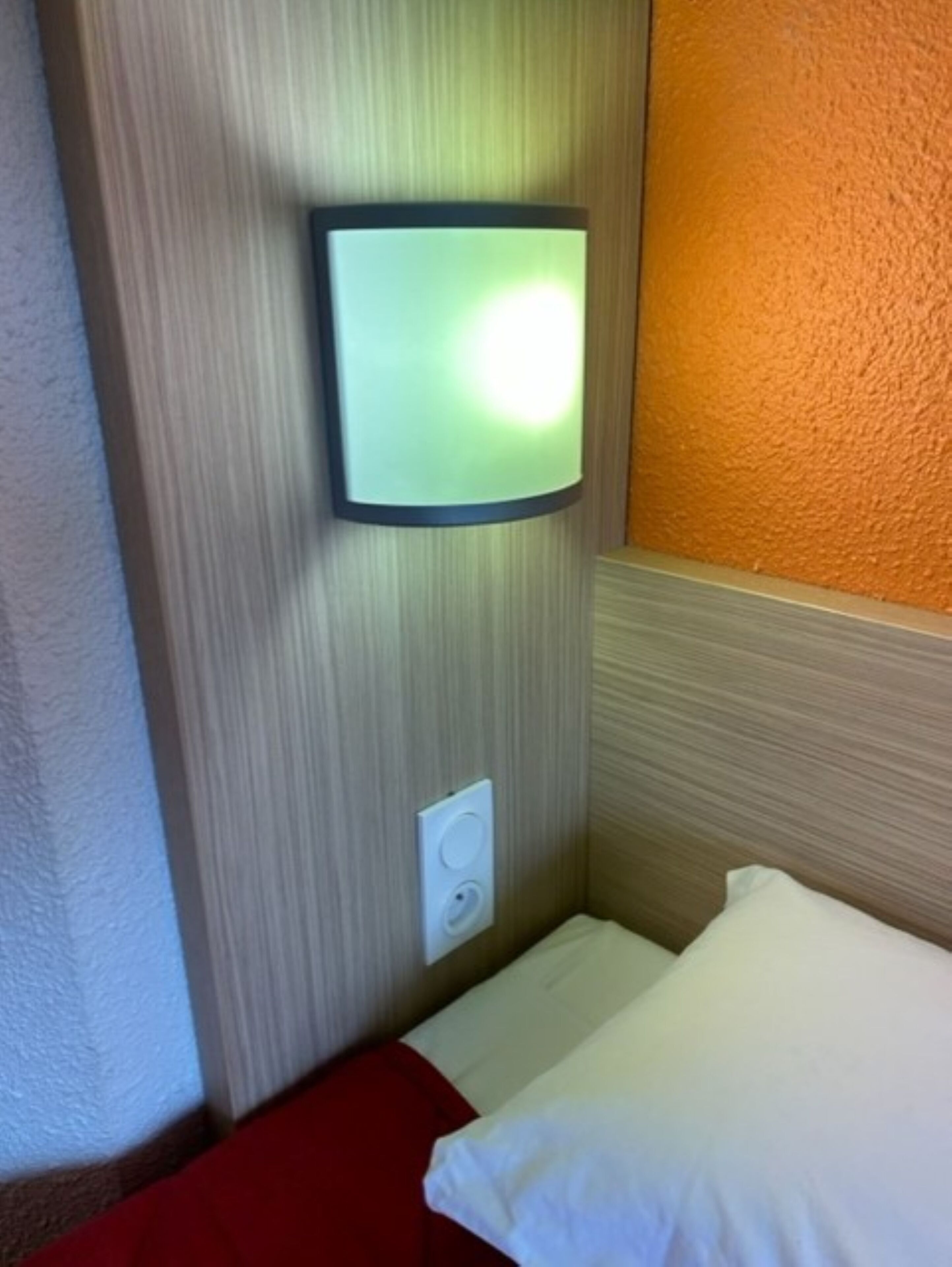 A hotel room with a bed, a wall-mounted light, and a power outlet.