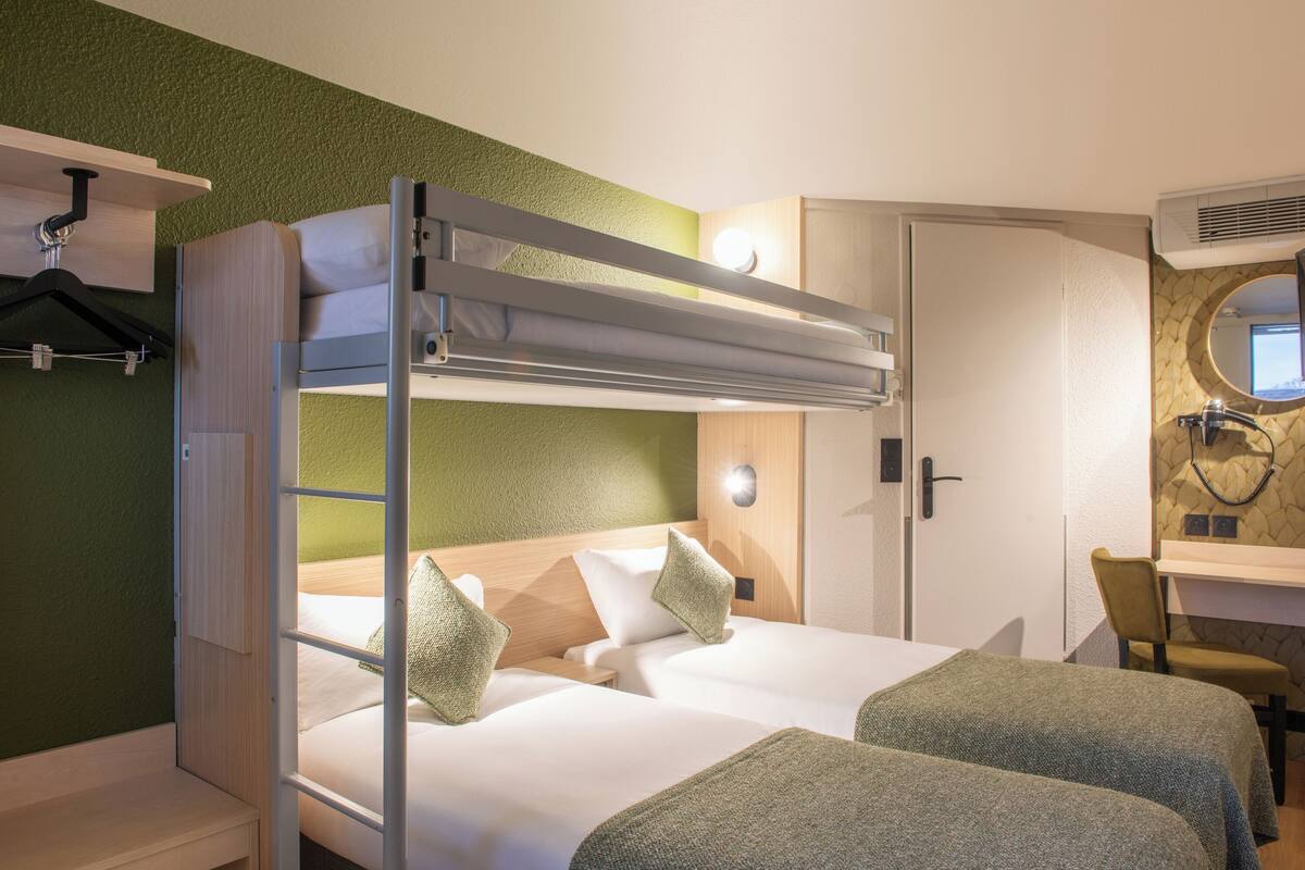 Une chambre d’hôtel avec un lit superposé, un bureau avec une chaise, un miroir et une petite salle de bain.
