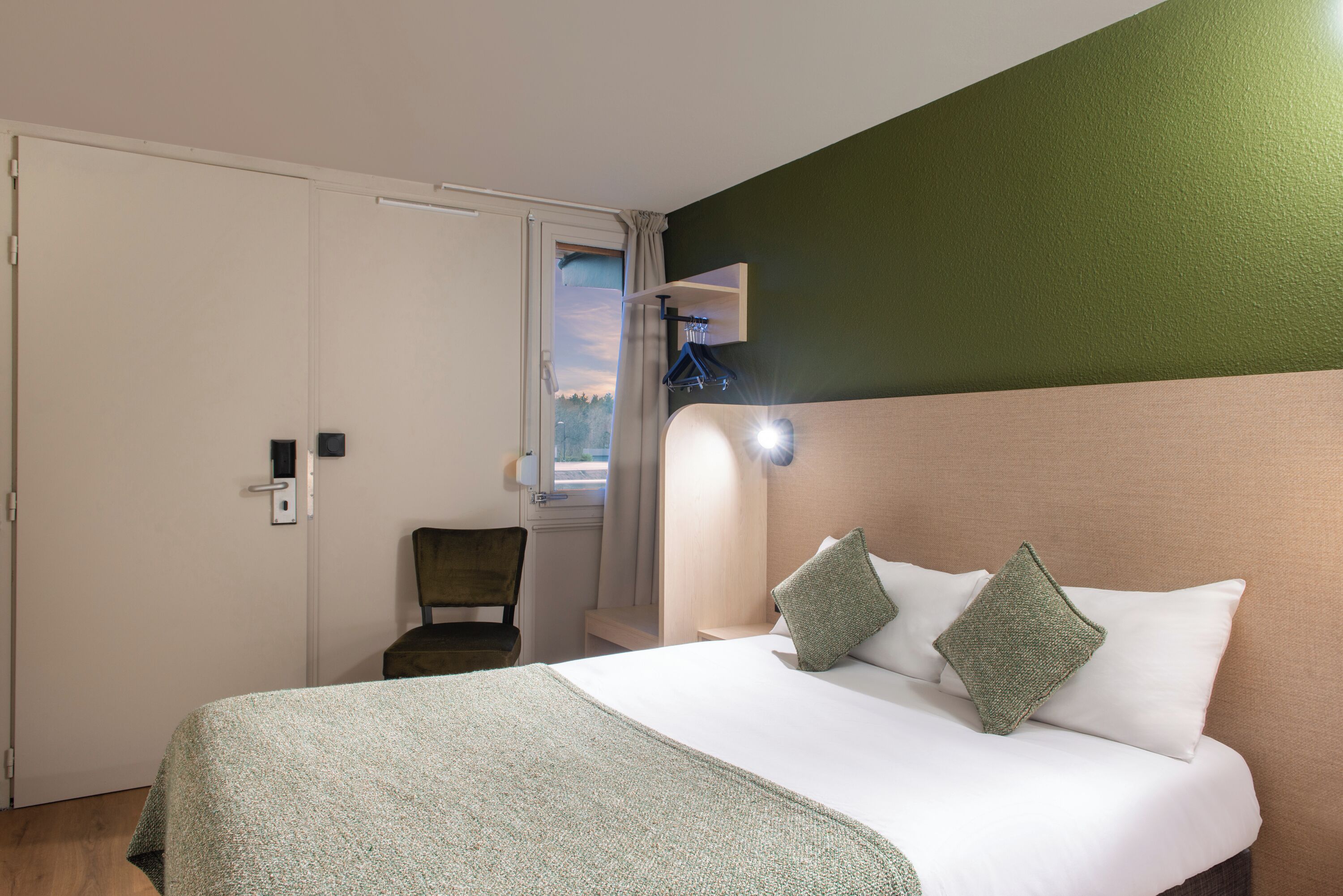 A hotel room with a bed, a chair, a window with curtains, and a wall-mounted light fixture.