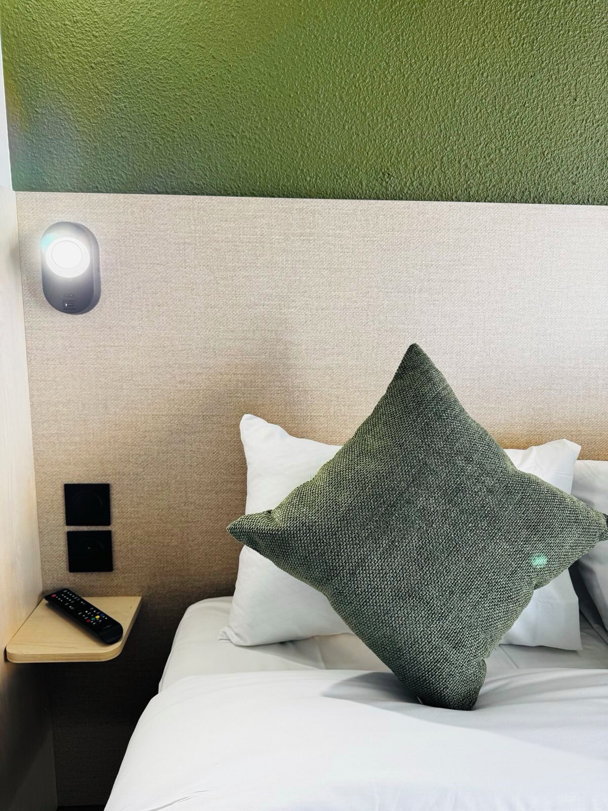 A hotel room with a bed, pillows, a remote control, and a wall-mounted light.