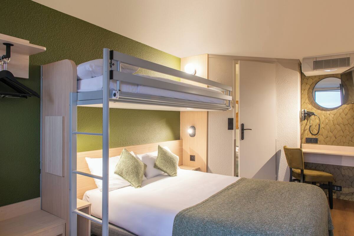 Un lit superposé avec un bureau et une chaise, une petite table de chevet et un miroir, dans une pièce aux murs verts et au plancher en bois.
