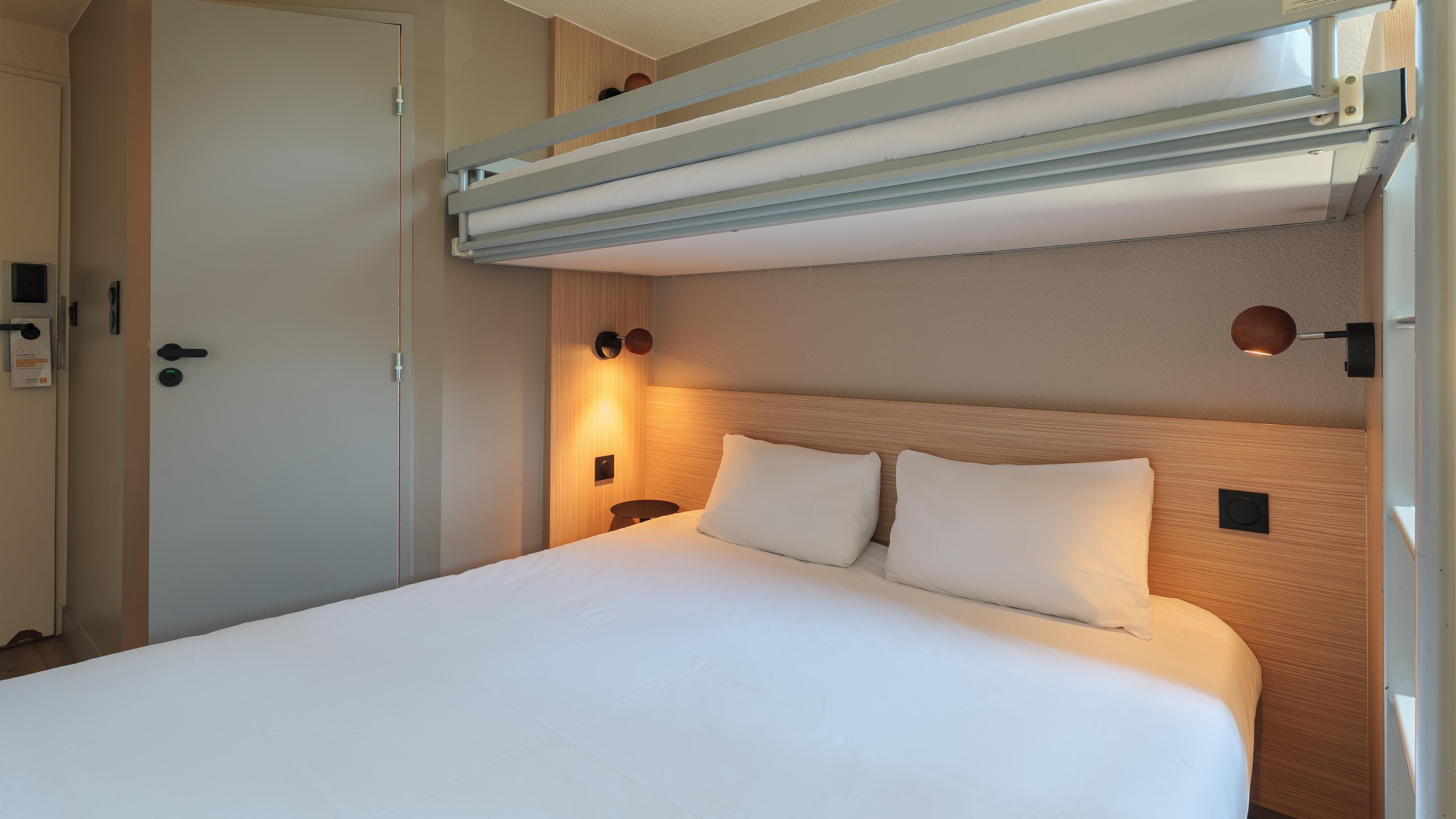 Een hotelkamer met een bed, een deur, een lamp en een bedieningspaneel aan de muur.