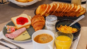 Een ontbijtplateau met toast, croissant, kaas, ham, boter, jam, koffie, cornflakes en sinaasappelsap.