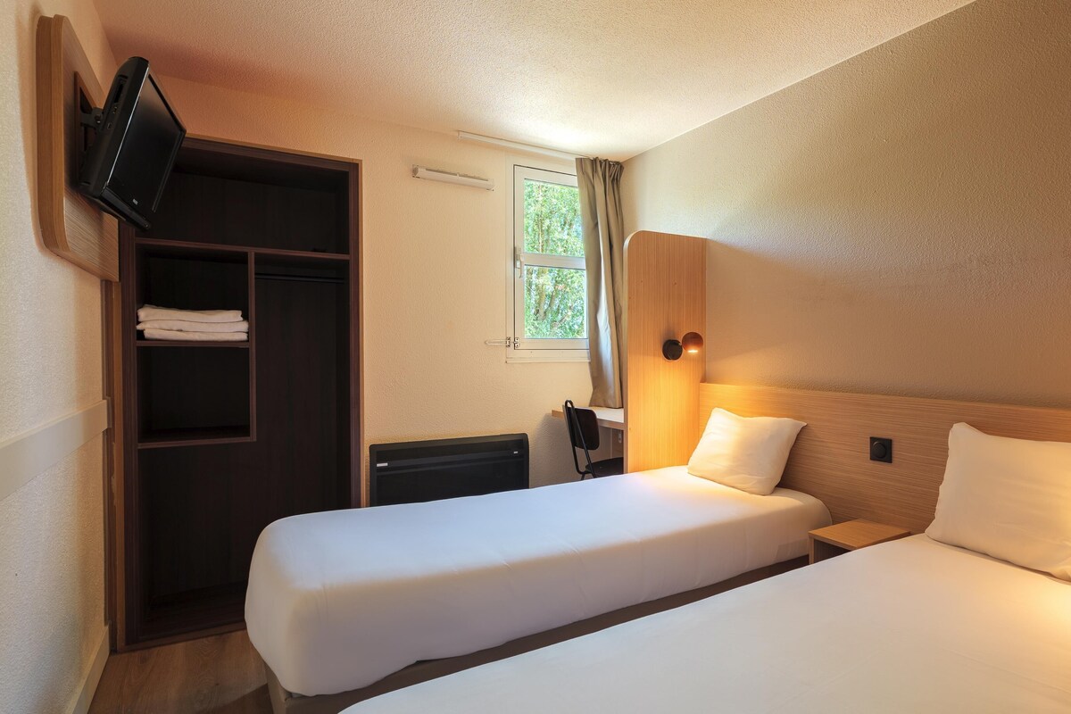 Ein Hotelzimmer mit zwei Betten, einem Schreibtisch, einem an der Wand montierten Fernseher und einem Fenster mit Vorhängen.